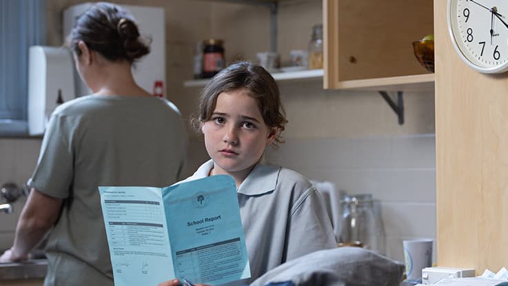 a girl reading her school report with her mum in the background in the kitchen