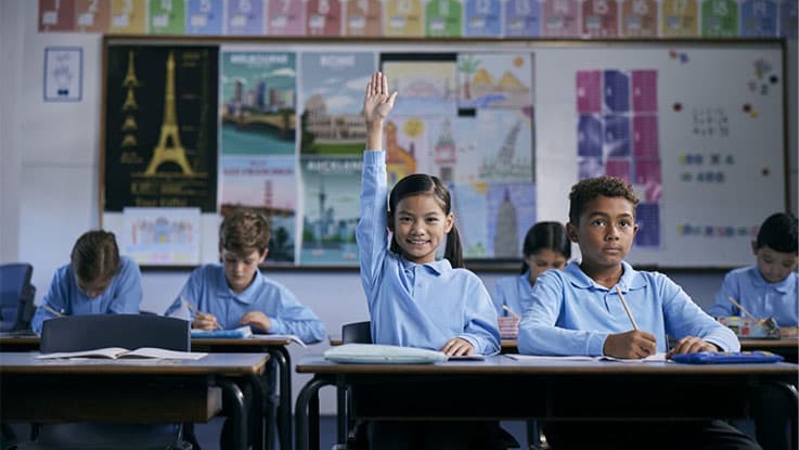 Girl sitting at classromm desk looking at camera with her hand up ready to answer a question