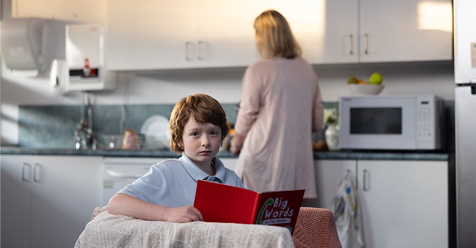 A young boy sitting at kitchen table reading with mum cooking in background