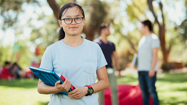 tertiary student outside holding a book and a laptop