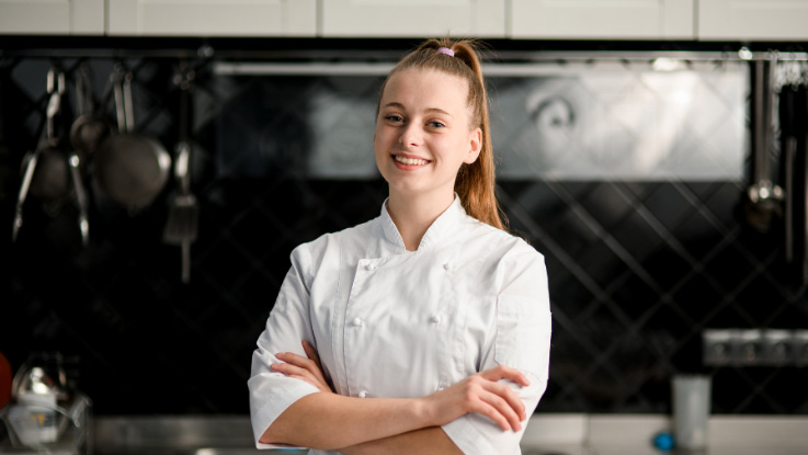 girl with light brown hair in a ponytail wearing a chefs jacket standing in front of a kitchen with her hands folded