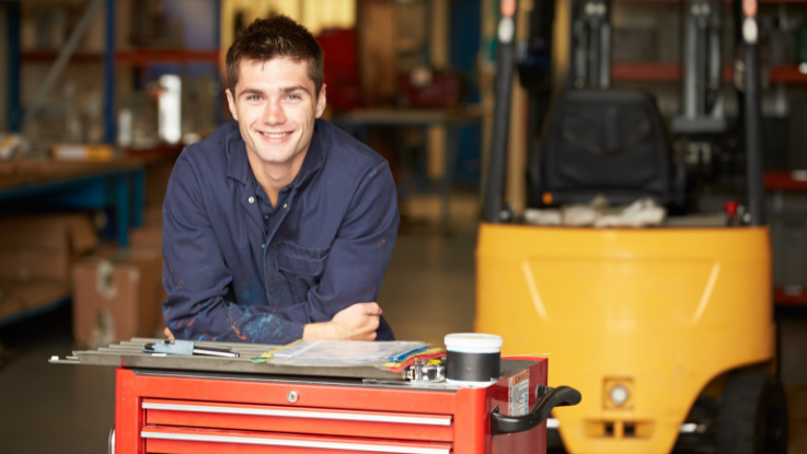 man with short brown hair wearing a blue mechanics uniform standing in a factory