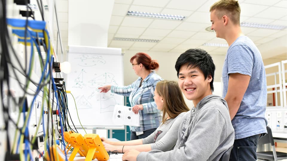 Students in a classroom working on electronics while a teacher explains at the whiteboard. A smiling student is in the front.