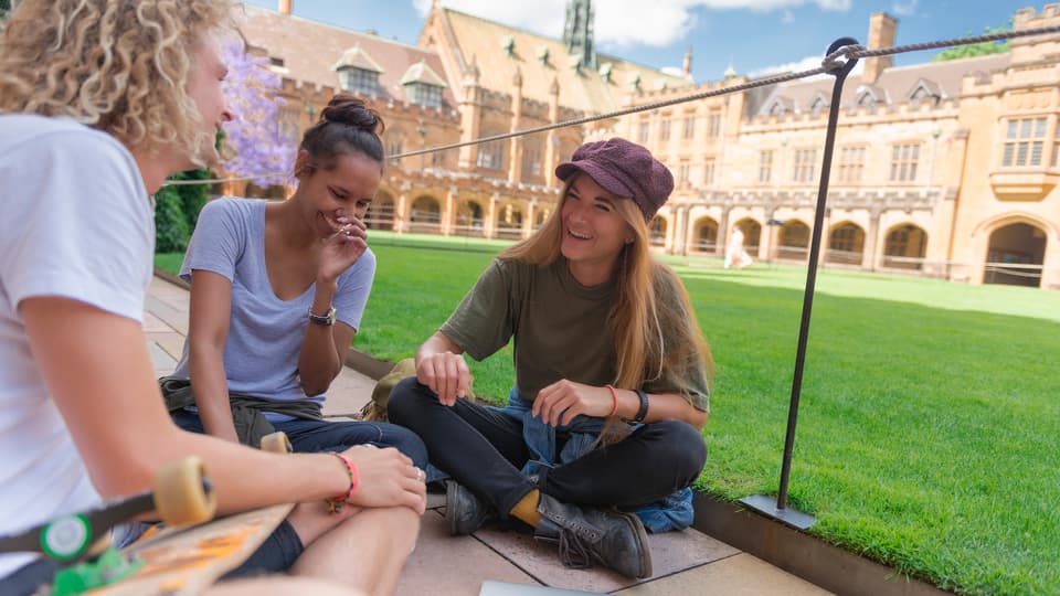 group of happy university students sitting on campus lawn, laughing and socializing, with historic college building in the background