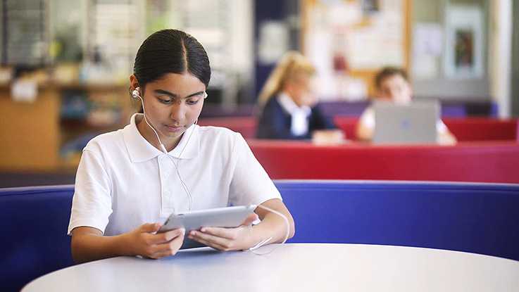 focused student in a white uniform using a tablet with earphones in a classroom, while other students work on laptops in the background