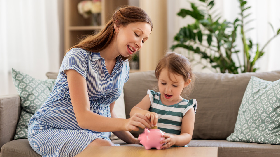 mother and daughter sitting on a couch, smiling while placing coins into a pink piggy bank, teaching kids about saving money at home.