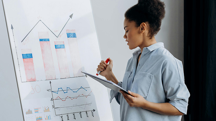 Woman holding a pen and clipboard looks at graphs on a whiteboard