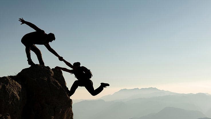 silhouette of one person holding hand for another person who is trying to climb up on cliff