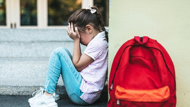 Sad small girl seating on floor with hand covering face and next to her there is read backpack 